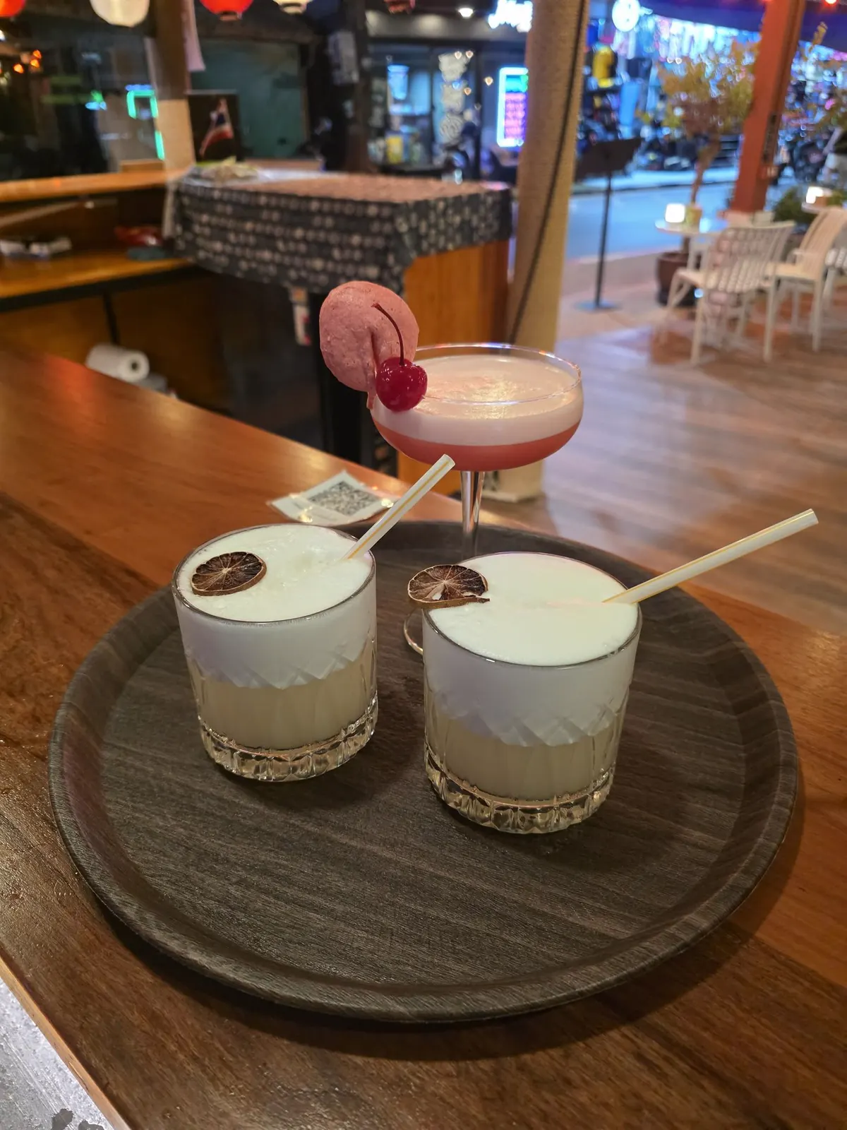 Whisky sours and a pink cocktail on a tray, the terrace and Lamai street behind at night